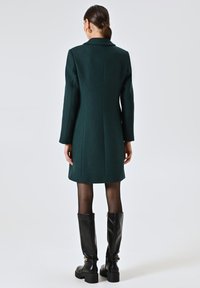 Donkergroene wollen jas met een getailleerde pasvorm, lange mouwen en een kraag, gecombineerd met kniehoog zwarte leren laarzen. Schoon, gestructureerde stoffen afwerking.