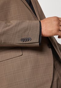 Brun rutig blazerärm med svart foder, försedda med fyra mörka knappar och en slät yta. Den slim fit-designen framhäver en skräddarsydd struktur.