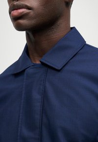 Close-up do rosto inferior e pescoço de uma pessoa a usar uma camisa ou jaqueta com padrão xadrez azul escuro contra um fundo simples.