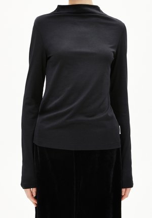 Femme portant un haut noir ajusté à manches longues avec un col haut légèrement drapé et un pantalon noir, sur un fond uni.