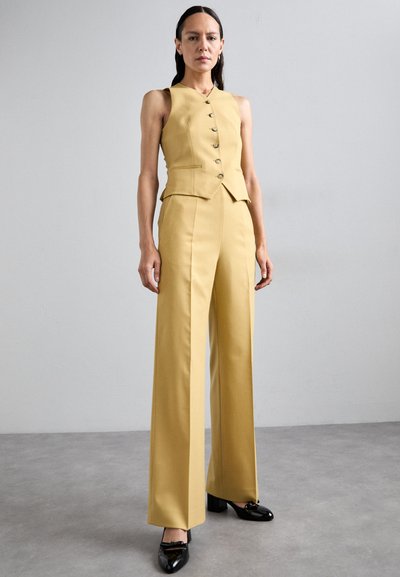 Gilet giallo con un colletto alto, cinque bottoni e vita a peplum, abbinato a pantaloni a gamba larga, entrambi realizzati in tessuto morbido. Tacchi neri con dettagli a fiocco.
