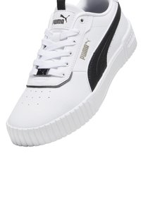Zapatilla blanca con acentos negros, parte superior sintética texturizada, suela de goma, puntera perforada y cordones. Presenta el logo de Puma en la lengüeta y el lateral.