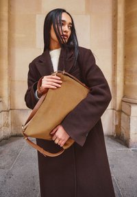 Brown wool coat over a light sweater, holding a tan canvas bag with brown leather strap and metal accents, standing against a stone wall.