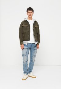 Young man standing against white wall wearing olive jacket over gray hoodie, white t-shirt, light blue cargo jeans, and white sneakers.