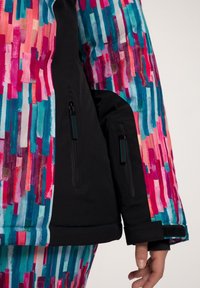 Giacca da sci colorata con vivaci strisce verticali in rosa, blu e viola, con dettagli neri e tasche con zip sulle maniche.