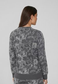 Vrouw met steil, donker haar, gekleed in een lange grijze sweatshirt met patroon en bijpassende broek, met de rug naar de camera tegen een effen achtergrond.