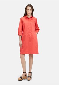 Korallenfarbendes Hemdkleid im klassischen Schnitt mit durchgehender Knopfleiste, Dreiviertelärmeln und glatter Textur. Kombiniert mit schwarzen Plateau-Sandalen.