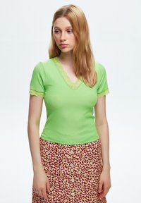 Top verde a coste con maniche corte, con pizzo al collo e alle maniche, abbinato a una gonna a bottoni con motivo in rosso e crema.