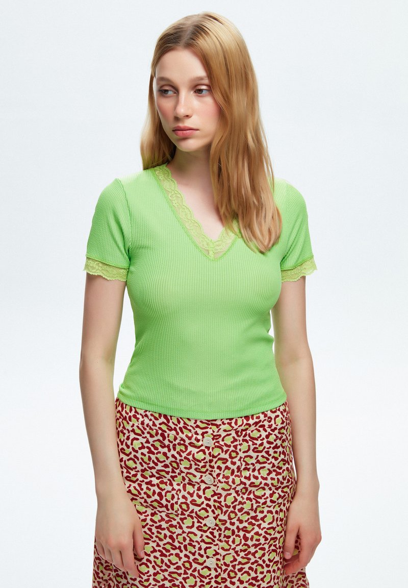 Top verde a coste con maniche corte, con pizzo al collo e alle maniche, abbinato a una gonna a bottoni con motivo in rosso e crema.