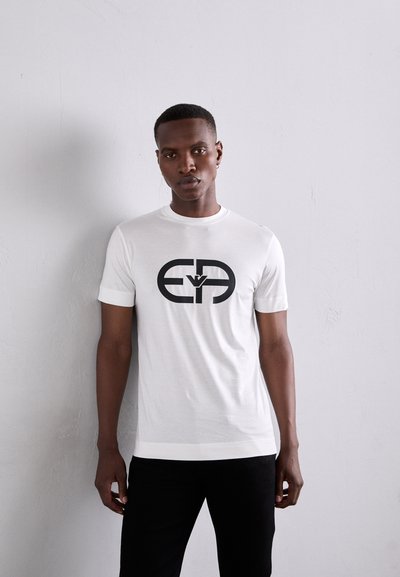 Tricou alb cu maneci scurte, din tricot jersey, având un mare logo grafic negru în centru. Designul are un guler rotund și o croială ajustată.