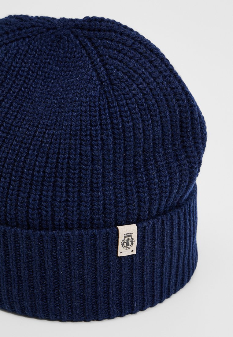 Granatowy, prążkowany czapka beanie z podwiniętym brzegiem i małą beżową metką z czarnym emblematem na białym tle.