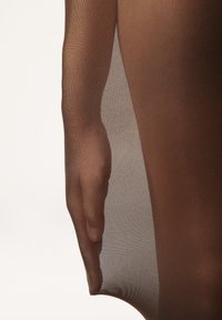 Des collants bruns transparents tombent sur une main, mettant en valeur une texture lisse avec un motif fin et côtelé. Le matériau paraît léger et délicat.
