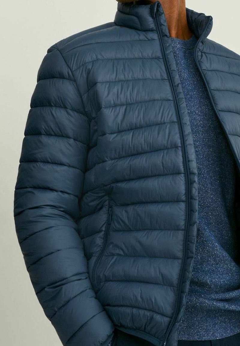 Veste matelassée bleu marine avec un design matelassé et un col montant, dotée de poches latérales zippées et d’une texture lisse et brillante.