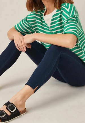 Vrouw zittend met armen rustend op knieën, draagt een groen-wit gestreept shirt, donkere skinny jeans en zwarte sandalen met gesp.