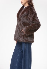Cappotto in pelliccia sintetica marrone con ampio colletto, tasche laterali e maniche lunghe. Tessuto texturizzato con un aspetto morbido e fluffy. Abbinato a jeans a gamba larga.
