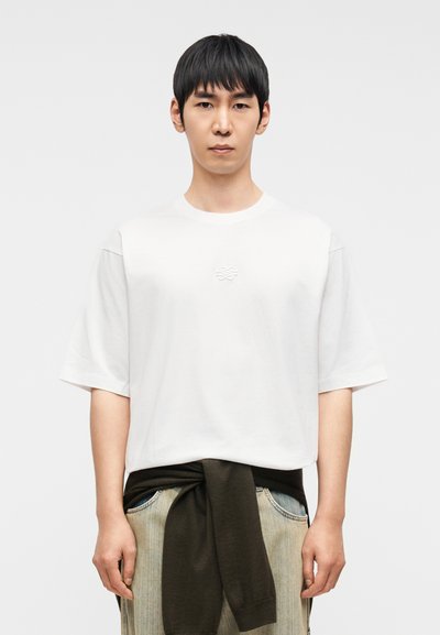 Λευκό t-shirt με κοντά μανίκια, στρογγυλή λαιμόκοψη και ανάγλυφο λογότυπο στο στήθος. Το μοντέλο το φοράει με σκούρο πράσινο πουλόβερ δεμένο γύρω από τη μέση.