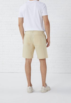 Homme portant un short beige, un t-shirt blanc et des baskets blanches, debout dos à la caméra contre un mur de briques blanches.