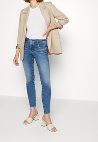 Kvinna klädd i beige blazer, vit topp, blå smala jeans och krämfärgade öppna klackar, stående med ena handen på höften mot en vit bakgrund.