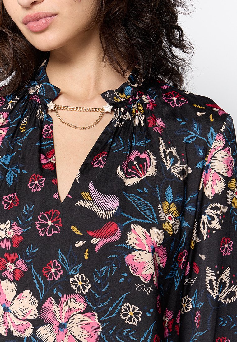 Blusa nera floreale con fiori ricamati colorati, scollatura a V profonda, colletto arricciato e un accento decorativo a catena sul collo.