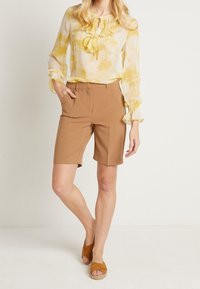 Gul blus med krage och volanger, i kombination med beige skräddarsydda shorts. Modellen bär bruna kilklackssandaler, vilket visar ett avslappnat sommaroutfit.