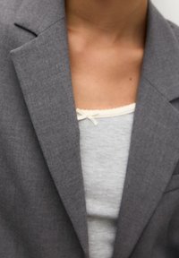 Gros plan d'une personne portant un blazer gris sur un haut gris clair avec un petit nœud blanc près de l'encolure.