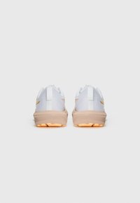 White athletic shoes with mesh upper, beige midsole, and rubber outsole. Back view shows contrasting yellow accents and heel tab.