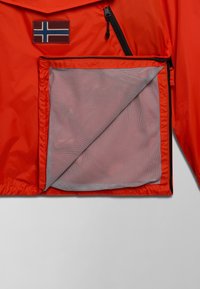 Oranje lichtgewicht jack met een netvak, diagonale ritssluiting en een patch met een blauw-wit vlagembleem. Gestructureerd materiaal.