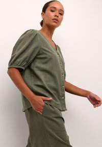 Blouse en lin vert avec des manches courtes bouffantes, décolleté en V et boutons sur le devant. Le modèle pose avec les mains dans les poches, mettant en valeur une coupe décontractée.