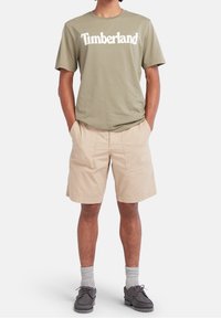 Olivengrøn kortærmet T-shirt med hvid "Timberland" tekst, parret med beige shorts, grå sokker og mørkegrå sko.