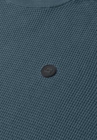 Tissu tricoté bleu texturé avec un patch circulaire en caoutchouc noir présentant un motif géométrique au centre.