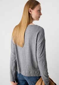 Kvinne med langt, rett, blondt hår, iført en grå langermet topp og blå jeans, holder en brun jakke i sin høyre hånd.