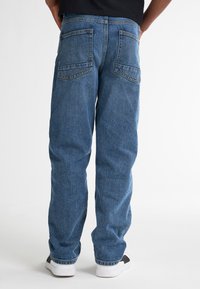 Jeans in denim blu con vestibilità rilassata, due tasche posteriori con cuciture dritte e una texture leggermente sbiadita. Indossati con sneakers bianche.