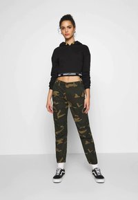 Sweatshirt cropped preta com detalhes amarelos, combinado com calças cargo em camuflagem verde. Modelo a usar ténis pretos e brancos. Conjunto urbano simples.
