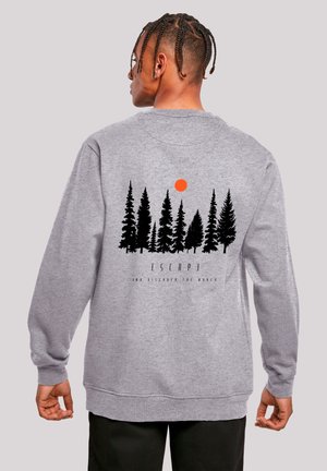 Mann mit geflochtenem Haar trägt grauen Pullover mit schwarzen Kiefern-Silhouetten und orangefarbenem Sonnenmotiv sowie dem Schriftzug "Escape" auf dem Rücken.