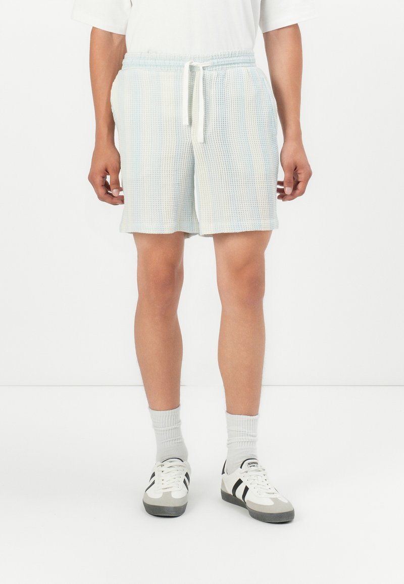 Pantalones cortos a cuadros en azul claro, confeccionados en una tela texturizada, con una cintura elástica y cordón ajustable, complementados con zapatillas blancas y calcetines grises.