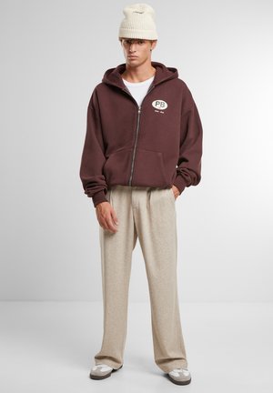Felpa con zip bordeaux con logo bianco, pantaloni ampi beige con texture, e berretto lavorato a maglia beige. Calzature casual con accenti bianchi e marroni.
