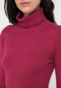 Εφαρμόζουσα μπλούζα με ζιβάγκο σε πλούσιο ροζ χρώμα, κατασκευασμένη από πλεκτό ύφασμα με ραβδώσεις και απαλή υφή, με ψηλό, διπλωμένο γιακά.