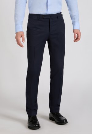 Mann trägt schlanke dunkelblaue Hose mit schwarzen Lederloafern und einem hellblauen Langarmhemd, steht auf einem grauen Boden.