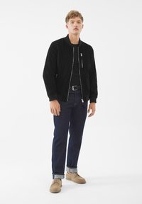 Svart bomberjacka med dragkedja, mörka jeans, beige skor och ett svart bälte, med en avslappnad passform och ribbade ärmslut. Enfärgad bakgrund.