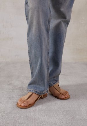 Person, die hellblaue, weit geschnittene Jeans und braune Zehensandalen trägt, steht auf einem hellgrauen Boden.