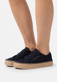 Mörkblå canvas sneakers med en vävd jutesula, med metallöglor, snörning och dekorativ sömnad runt sulans kant.