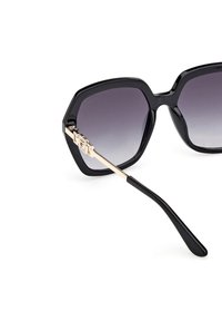 Zwarte hexagonale zonnebril met verlopende glazen, gouden accenten op de pootjes en een glanzende afwerking. Getextureerde zijdetails nabij de scharnieren.