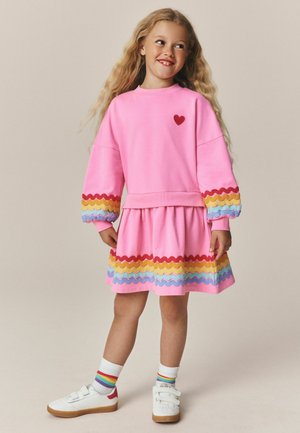 Niña joven con un vestido rosa con rayas onduladas de arcoíris, zapatillas blancas y calcetines con rayas multicolor, de pie y sonriendo.