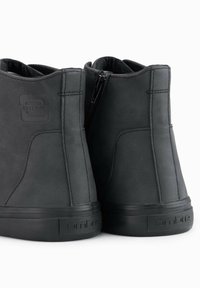 Botas de cuero negro hasta el tobillo con una textura suave, con cremallera lateral y suelas de goma. Logo grabado en el tacón.