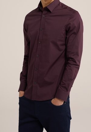 Uomo che indossa una camicia a maniche lunghe color viola scuro con bottoni, con entrambe le mani nelle tasche dei pantaloni blu scuro, davanti a uno sfondo semplice.