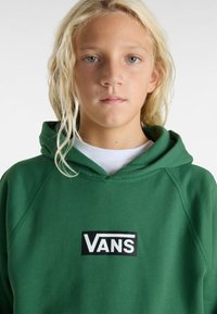 Sudadera verde hecha de una tela suave con un bolsillo tipo canguro en el frente, con un logo negro de Vans sobre un fondo blanco.