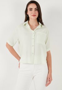 Cameria verde chiaro con maniche corte, camicia con bottoni e tasca, realizzata in un tessuto morbido e texturizzato, caratterizzata da un design rilassato e corto.