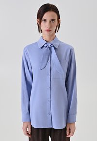 Camicia azzurra chiara con maniche lunghe, dettaglio con cravatta al colletto e una piccola tasca sul petto. Tessuto liscio con una leggera lucentezza.