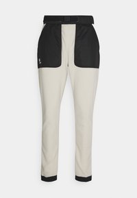 Byxor i mjukt tyg i ljusbeige färg med svarta nylonpaneler vid höfterna. Har en midja med bälte och åtsittande muddar.