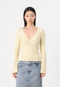 Lace långärmad topp i ljusgult med V-ringning, som har intrikata blommönster och en figursydd silhuett, parat med denim.